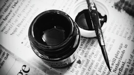 A close-up of an open bottle of black ink, accompanied by a paintbrush and a white bottle cap sitting on a newspaper page with visible text.