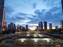 A cityscape featuring a modern skyline with numerous high-rise buildings under a dramatic evening sky. The foreground includes a construction site with visible structures and equipment. The buildings are illuminated, indicating the onset of nightfall.