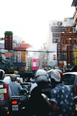 A bustling urban street scene filled with various brightly colored signs featuring different languages, likely in an Asian city. Vehicles and people on scooters are visible in the foreground, while multiple store signs and banners are layered densely in the background, creating a vibrant cityscape.