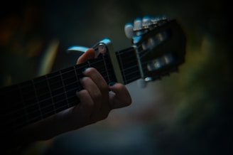 A close-up of a hand playing an acoustic guitar with fingers pressing down on the strings and frets. The background is blurred, creating a soft focus effect, while the lighting contributes to a moody atmosphere.