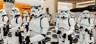 A group of people dressed in white and black futuristic armor, resembling characters from a popular sci-fi franchise, are standing in a formation. They are inside a well-lit commercial space with orange-lit glass and signage displayed in the background.