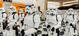 A group of people dressed in white and black futuristic armor, resembling characters from a popular sci-fi franchise, are standing in a formation. They are inside a well-lit commercial space with orange-lit glass and signage displayed in the background.