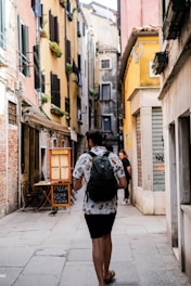 A narrow European street lined with colorful buildings, featuring a man walking away with a backpack. The walls of the buildings have shutters and small planters with greenery. A chalkboard sign outside advertises pizza, creating a quaint and inviting atmosphere.