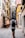 A narrow European street lined with colorful buildings, featuring a man walking away with a backpack. The walls of the buildings have shutters and small planters with greenery. A chalkboard sign outside advertises pizza, creating a quaint and inviting atmosphere.