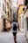 A narrow European street lined with colorful buildings, featuring a man walking away with a backpack. The walls of the buildings have shutters and small planters with greenery. A chalkboard sign outside advertises pizza, creating a quaint and inviting atmosphere.