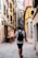 A narrow European street lined with colorful buildings, featuring a man walking away with a backpack. The walls of the buildings have shutters and small planters with greenery. A chalkboard sign outside advertises pizza, creating a quaint and inviting atmosphere.