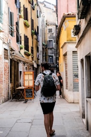 A narrow European street lined with colorful buildings, featuring a man walking away with a backpack. The walls of the buildings have shutters and small planters with greenery. A chalkboard sign outside advertises pizza, creating a quaint and inviting atmosphere.