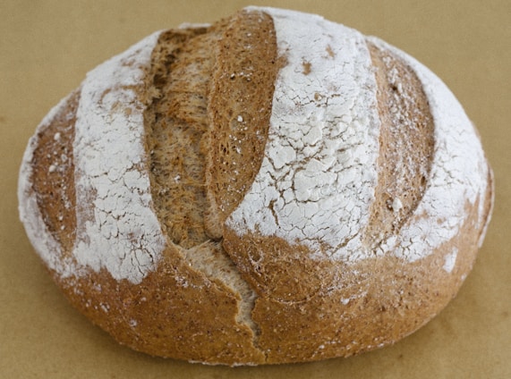 A round loaf of artisan bread with a cracked, crusty surface and a pattern of flour dusted over the top. The bread has a rustic appearance with a golden-brown color and deep slashes in the crust.