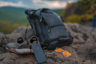 On a rocky surface, a pair of worn-out slip-on shoes are placed next to a black backpack. A smartphone rests with its screen visible, showing the time as 5:43. Nearby, orange-tinted sunglasses add a pop of color to the scene. In the background, an out-of-focus landscape of lush green hills stretches out under a cloudy sky.