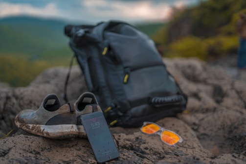 On a rocky surface, a pair of worn-out slip-on shoes are placed next to a black backpack. A smartphone rests with its screen visible, showing the time as 5:43. Nearby, orange-tinted sunglasses add a pop of color to the scene. In the background, an out-of-focus landscape of lush green hills stretches out under a cloudy sky.