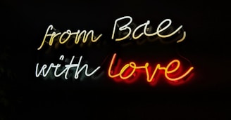 Neon sign displaying the text 'from Bae, with love' in cursive script against a dark background. The words are illuminated in different colors, creating a striking contrast with the darkness surrounding it.