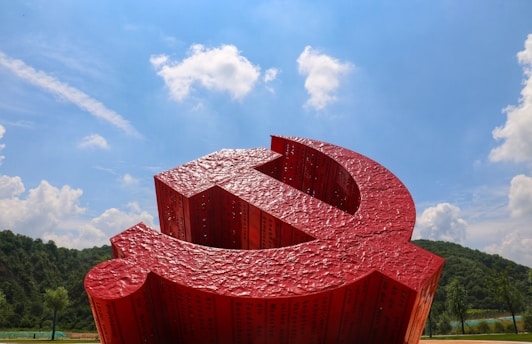 A large red sculpture resembling a hammer and sickle is prominently displayed against a backdrop of a clear blue sky with scattered white clouds. The sculpture has a textured surface and appears to be part of a landscaped area with green hills and trees in the background.