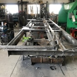 A workshop setting with a partially assembled metal frame on a workbench. Various tools and equipment, including a welding machine, are scattered around. Industrial shelving holds miscellaneous items in the background, and gloves rest on the frame indicating ongoing work.