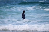 A person wearing a dark wetsuit stands waist-deep in the ocean, surrounded by waves. The water is a mix of blue and white, indicating movement and energy.