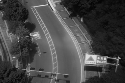 A curved road is bordered by dense trees on one side, with a crosswalk and traffic cone visible. A traffic sign with Korean text is placed prominently at the curve. The road's surface is mostly clear, with a slight shadow from nearby foliage.