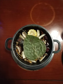 A metal pot filled with a variety of ingredients including mushrooms, corn, and herbs. A large green leaf with writing is placed on top of the contents. The pot is set on a wooden table, adding a rustic feel.