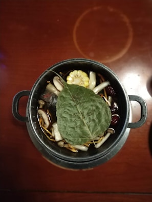 A metal pot filled with a variety of ingredients including mushrooms, corn, and herbs. A large green leaf with writing is placed on top of the contents. The pot is set on a wooden table, adding a rustic feel.