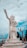 A large white statue of a guardian or deity with intricate detailing stands on a tiered pedestal. The figure is pointing upwards with its right hand and has an ornate headdress and armor. Surrounding the statue are bare tree branches and a coastal or temple backdrop under a partly cloudy sky.