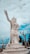 A large white statue of a guardian or deity with intricate detailing stands on a tiered pedestal. The figure is pointing upwards with its right hand and has an ornate headdress and armor. Surrounding the statue are bare tree branches and a coastal or temple backdrop under a partly cloudy sky.