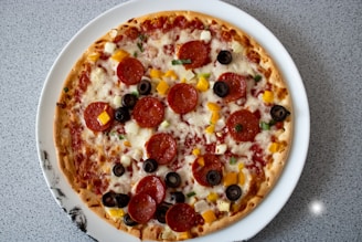 A round pizza topped with pepperoni slices, black olives, diced yellow bell peppers, and onions, placed on a white plate. The pizza is prepared on a thin crust with melted cheese and a layer of tomato sauce. The background consists of a speckled gray countertop.
