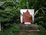 A terracotta relief sculpture is set against a white backdrop in a lush garden. The sculpture depicts a seated figure holding another figure lying across their lap, reminiscent of a religious or classical theme. Surrounding the sculpture are dense green plants and trees, contributing to a serene and natural atmosphere.