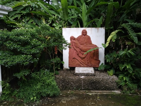 A terracotta relief sculpture is set against a white backdrop in a lush garden. The sculpture depicts a seated figure holding another figure lying across their lap, reminiscent of a religious or classical theme. Surrounding the sculpture are dense green plants and trees, contributing to a serene and natural atmosphere.