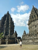 Intricate stone temple structures rise dramatically against a backdrop of a bright blue sky with sparse white clouds. A person stands in the foreground, wearing casual tourist attire. Lush greenery and stone pathways add to the ancient architectural setting.