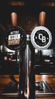 Two wooden-handled taps labeled 'Nitro Cold Brew' and 'CB Cold Brew' are affixed to a shiny metallic dispenser. The background is dimly lit, with a warm wooden texture and blurred elements.