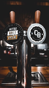 Two wooden-handled taps labeled 'Nitro Cold Brew' and 'CB Cold Brew' are affixed to a shiny metallic dispenser. The background is dimly lit, with a warm wooden texture and blurred elements.