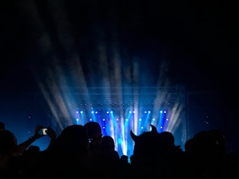 A concert stage with bright blue and white spotlights shining into the night sky, creating beams of light. Silhouettes of people in the audience are visible, with one person holding a phone to take a picture. The crowd is immersed in the atmosphere of the live performance.