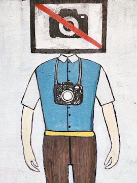 A painted figure with a rectangular box as its head, displaying a camera with a red diagonal line crossing it out. The figure is wearing a blue vest over a white shirt and brown pants, with a camera hanging from a strap around its neck.