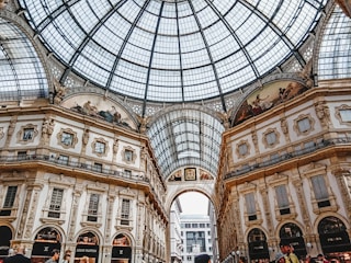 An ornate indoor shopping arcade with a grand glass dome ceiling and detailed architectural features. The walls have intricate moldings and frescoes, with luxury brand store signs visible at ground level. The structure is bustling with people, adding to the lively atmosphere.