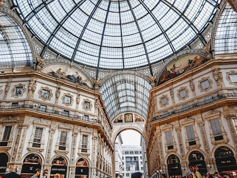 An ornate indoor shopping arcade with a grand glass dome ceiling and detailed architectural features. The walls have intricate moldings and frescoes, with luxury brand store signs visible at ground level. The structure is bustling with people, adding to the lively atmosphere.