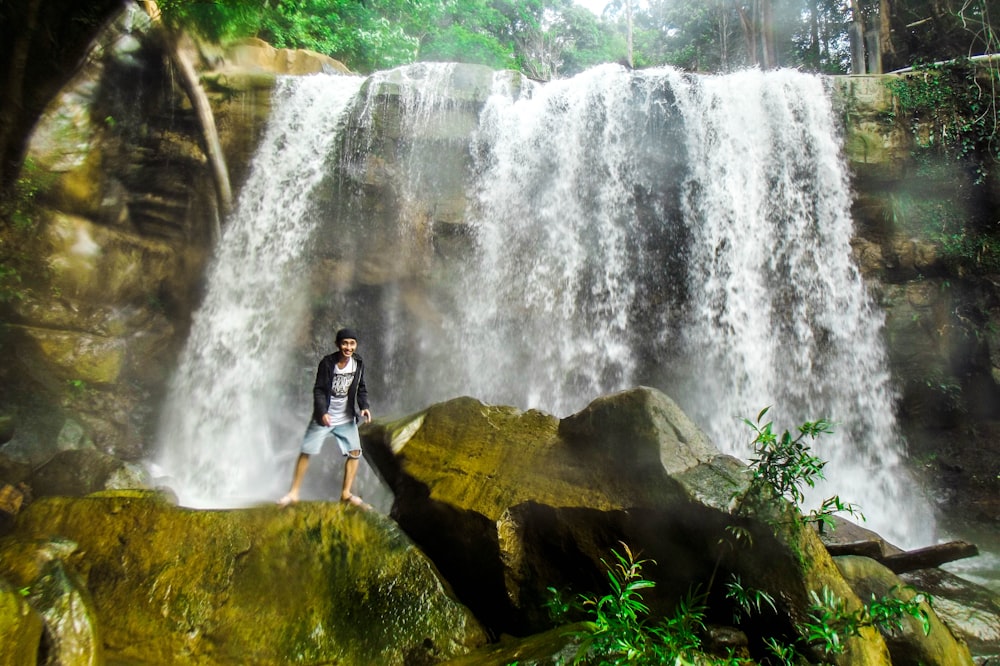 man standing near waterfalls surrounded with tall and green trees