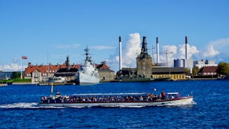 A tour boat filled with passengers travels on a body of water. In the background, a large ship is docked near several industrial buildings and structures with smokestacks. A Danish flag is visible beside traditional buildings with red roofs. The sky is clear with a few clouds.