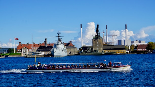 A tour boat filled with passengers travels on a body of water. In the background, a large ship is docked near several industrial buildings and structures with smokestacks. A Danish flag is visible beside traditional buildings with red roofs. The sky is clear with a few clouds.