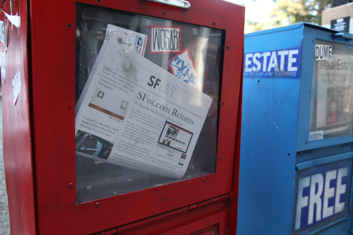 A newspaper vending machine painted red and containing a visible newspaper titled 'SFist' is partly obscured by a sticker. The machine is positioned next to a blue vending machine labeled 'Free Real Estate'. Both machines have a slightly worn appearance, with scratches and minor damage.