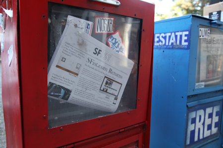 A newspaper vending machine painted red and containing a visible newspaper titled 'SFist' is partly obscured by a sticker. The machine is positioned next to a blue vending machine labeled 'Free Real Estate'. Both machines have a slightly worn appearance, with scratches and minor damage.