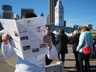 A person stands in an outdoor market area, holding and reading a newspaper with the headline 'SFist.com Returns.' Several people are walking or standing nearby, dressed in casual winter clothing. In the background, there is a prominent clock tower and a skyline of tall buildings under a clear blue sky.