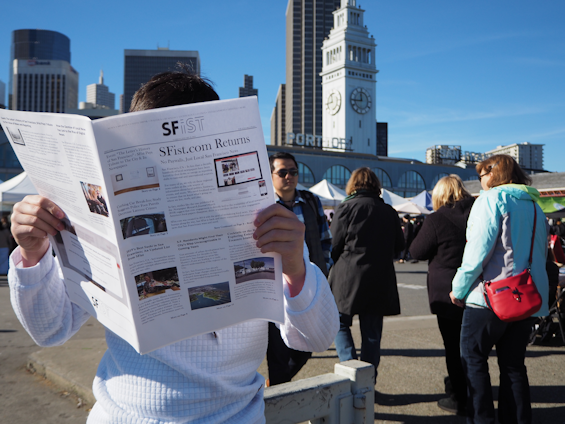 A person stands in an outdoor market area, holding and reading a newspaper with the headline 'SFist.com Returns.' Several people are walking or standing nearby, dressed in casual winter clothing. In the background, there is a prominent clock tower and a skyline of tall buildings under a clear blue sky.