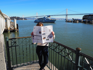 A person is standing by a waterfront railing, reading a newspaper that obscures their face. Behind them is a large body of water with a ferry passing by and a large suspension bridge further in the background. The scene includes clear blue skies and is brightly lit by sunlight.
