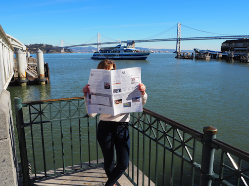 A person is standing by a waterfront railing, reading a newspaper that obscures their face. Behind them is a large body of water with a ferry passing by and a large suspension bridge further in the background. The scene includes clear blue skies and is brightly lit by sunlight.