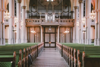 The interior of a large, ornate church with tall columns, green pews, hanging chandeliers, and an impressive pipe organ in the background. The center aisle is carpeted and leads directly to a set of wooden doors under the organ balcony.