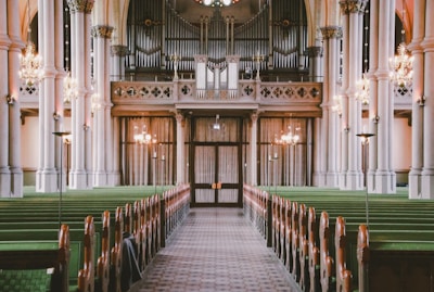 The interior of a large, ornate church with tall columns, green pews, hanging chandeliers, and an impressive pipe organ in the background. The center aisle is carpeted and leads directly to a set of wooden doors under the organ balcony.