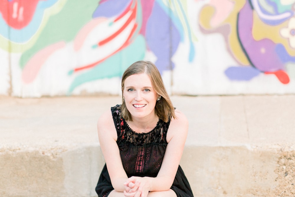 smiling woman wearing black sleeveless dress sitting in front of graffiti