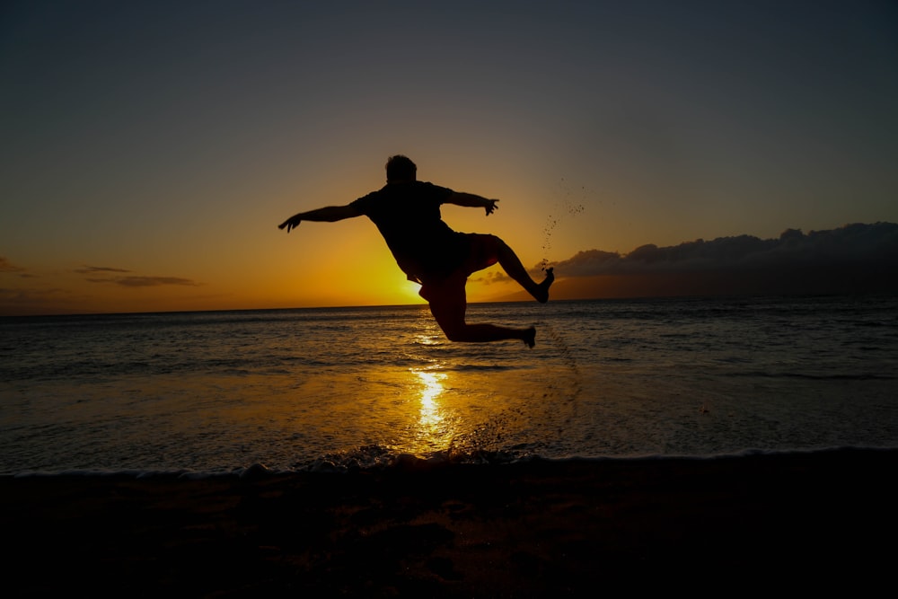 silhouette of jumping man on seashore during golden hour