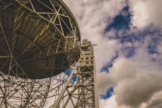 A massive radio telescope structure with intricate metallic framework is set against a dynamic sky filled with clouds. The perspective captures the underside of the dish and a portion of the supporting tower, emphasizing the industrial design and scale of the telescope.