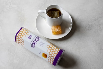 A white cup filled with tea sits on a matching saucer. Next to the cup is a piece of honeycomb. A rectangular package labeled 'honeycomb' is also visible on the table surface, which has a light, textured appearance.
