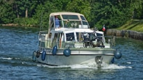 A boat is traveling on a calm body of water surrounded by greenery. On the deck, there is a bicycle. A person is visible at the helm under a canopy. Several fenders are hanging from the sides of the boat, and a flag is mounted at the rear.