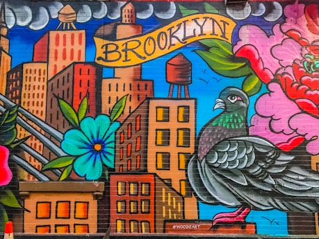 A vibrant mural depicting urban life with stylized skyscrapers and a large pigeon in the foreground. The word 'Brooklyn' is painted on a banner above the buildings. Bright flowers and clouds surround the scene, adding a lively and colorful backdrop.
